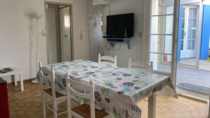 Een eethoek met een tafel, gedekt met een tafelkleed met dessin, witte stoelen, een aan de muur bevestigde televisie en een deur die naar buiten leidt.