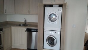 Eine kompakte Küche mit Holzausstattung, einem Edelstahl-Spülbecken, einem Geschirrspüler sowie einer übereinander angeordneten Waschmaschine und einem Trockner.