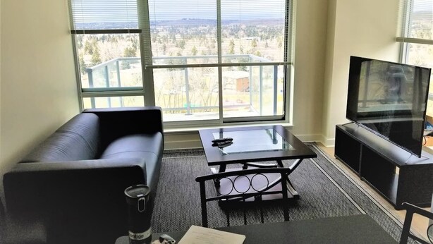 Ein modernes Wohnzimmer mit einem schwarzen Ledersofa, einem Glastisch und einem Flachbildfernseher auf einem schwarzen Standfuß. Große Fenster bieten Blick auf eine Stadtlandschaft.