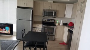 Eine kompakte Küche mit hellen Holzschränken, einem Edelstahlkühlschrank, einer Mikrowelle, einem Backofen und einem Geschirrspüler. Ein schwarzer Esstisch bietet Platz für vier Stühle.