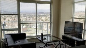 Ein modernes Wohnzimmer mit einer Couch, einem Glastisch, einem Fernseher und Blick auf eine Stadtlandschaft durch große Fenster.