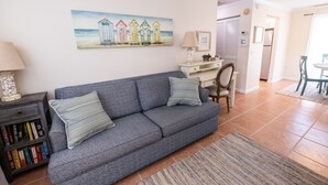 A living room with a grey sofa, a bookshelf, a painting of beach huts, and a dining area in the background. - LOCATION!! Cabana just steps from the beach! (Holmes Beach)