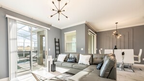 A spacious living room with a sectional sofa, a dining area with white chairs, and a large sliding glass door leading to an outdoor area.