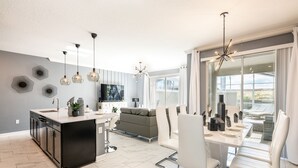 A modern kitchen with a central island, a dining area with white chairs, and a large window offering a view.