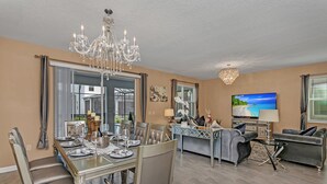 A dining area with a chandelier, a TV in the living room, and a large window with curtains. - Elegant 9 BR Mansion Center of Attractions 4515 (Kissimmee)