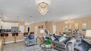 A spacious living room with a grey sofa set, a chandelier, and a dining area with a staircase. - Elegant 9 BR Mansion Center of Attractions 4515 (Kissimmee)