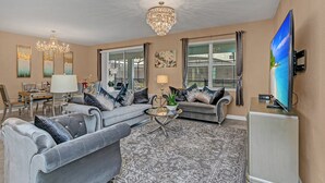 A living room with a grey sofa, a coffee table, a chandelier, and a television. - Elegant 9 BR Mansion Center of Attractions 4515 (Kissimmee)