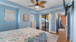 Ein Schlafzimmer mit einem blumigen Bettbezug, einem Deckenventilator, einer Glasschiebetür zu einem Balkon und einer weißen Kommode.
