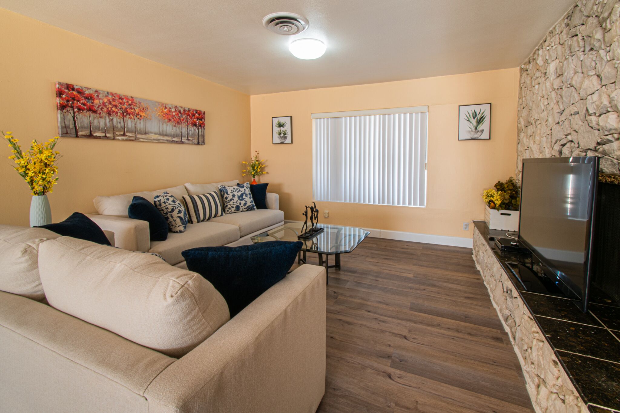 A living room with a beige sofa, a glass coffee table, a stone accent wall, a TV, and a window with blinds.