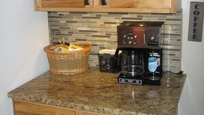 A kitchen with wooden cabinets, a coffee maker, a basket with kitchen utensils, and a coffee sign on the wall. - Steps away from Wild Basin:RMNP- 2-bdrm cabin perfect for adventure or relaxing! (Allenspark)