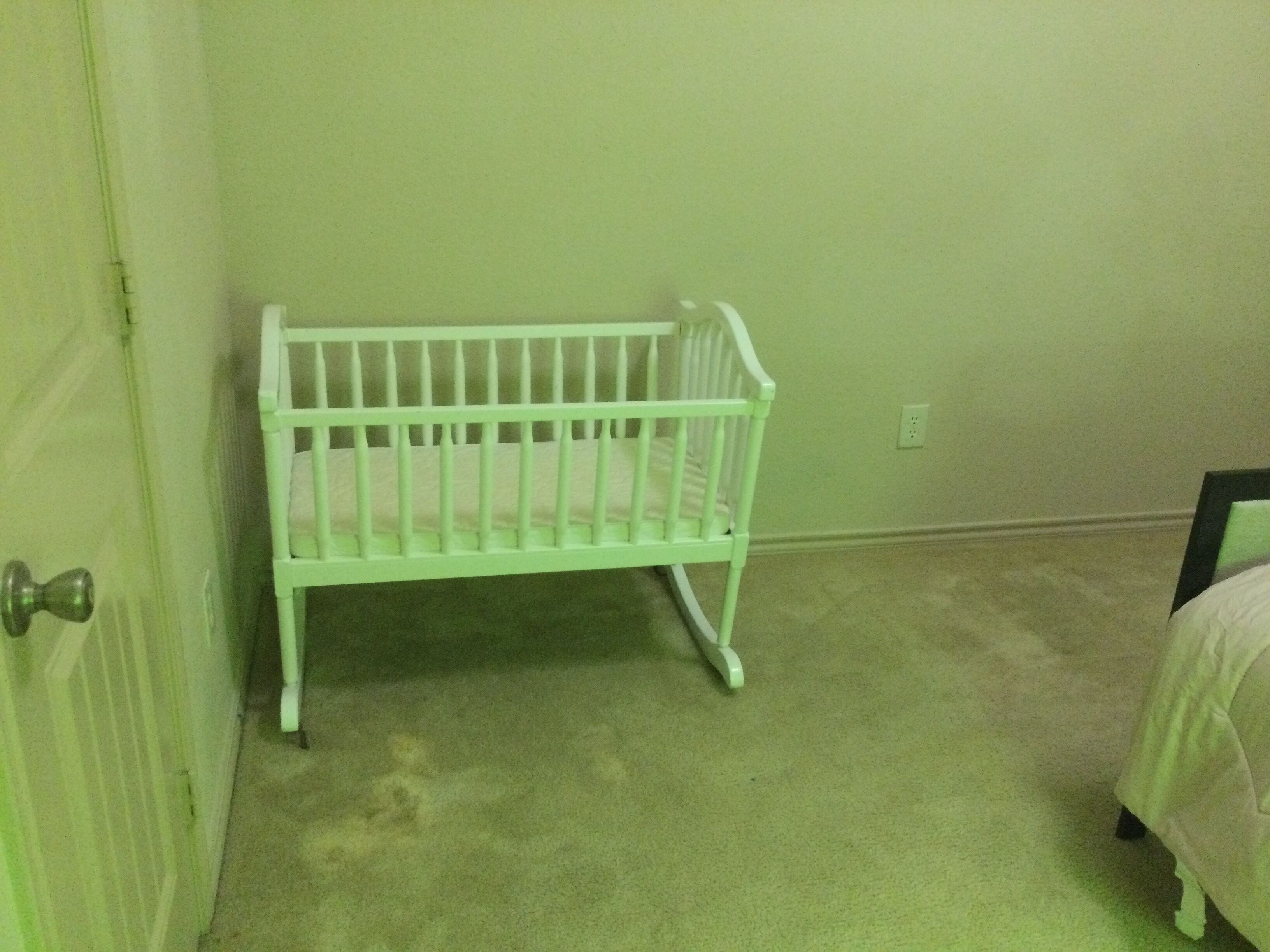 Una habitación con una cuna blanca, pared verde, suelo alfombrado y una puerta con picaporte plateado.