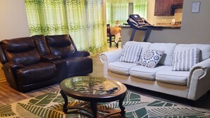 Uma sala de estar com um sofá de couro marrom, um sofá branco com almofadas estampadas, uma mesa de centro de vidro e um tapete grande com padrão verde e bege.