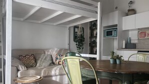 Un coin repas avec une table en bois, des chaises vertes, un canapé beige agrémenté de coussins à motifs et un petit tabouret.
