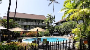 A pool area with lounge chairs and umbrellas, surrounded by lush greenery and a building with a balcony. - Experience historic charm near Kailua Pier - Walk to restaurants, shops,AC,Wifi (Kailua-Kona)
