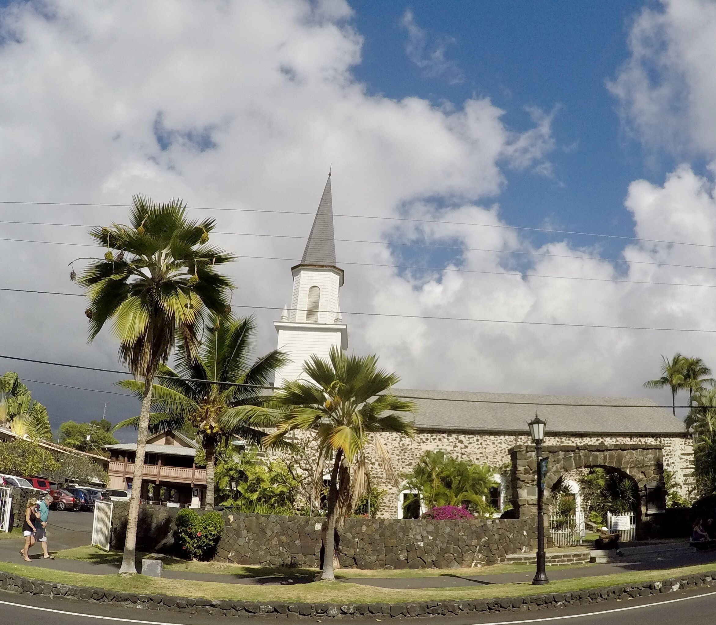 A church with a steeple, palm trees, and a stone wall.