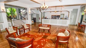 A spacious living area with wooden flooring, a large floral rug, and several wooden chairs and sofas. A staircase is visible in the background leading to another level. - Experience historic charm near Kailua Pier - Walk to restaurants, shops,AC,Wifi (Kailua-Kona)