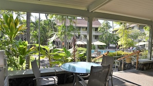 A covered outdoor seating area with a round table and chairs, surrounded by tropical plants and overlooking a pool and buildings. - Experience historic charm near Kailua Pier - Walk to restaurants, shops,AC,Wifi (Kailua-Kona)