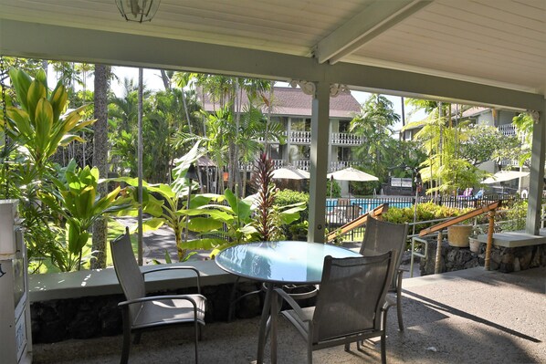 An outdoor seating area with a table and chairs, surrounded by tropical plants and a view of a pool and buildings.
