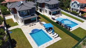 Et boligområde med to swimmingpools, hver omgivet af liggestole og parasoller, grønne plæner og huse med grå tage.