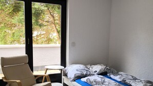 Habitació petita amb llit, cadira, taula i finestra amb vista a vegetació.