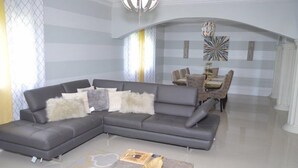 A modern living room with a grey sectional sofa, a coffee table, and a dining area with a chandelier. - Marvettes Hideaway (Lucea)