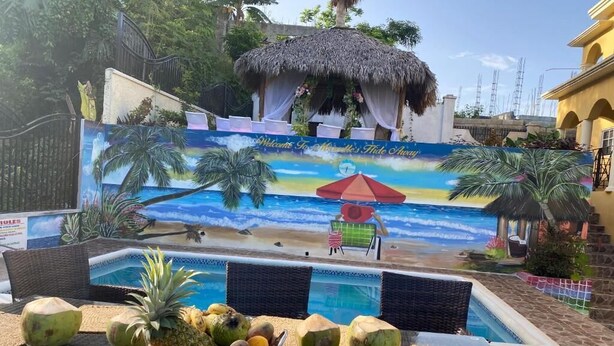 A poolside area with a tropical mural, a thatched-roof structure, and a table with coconuts and fruits. - Marvettes Hideaway (Lucea)