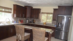 A kitchen with dark wooden cabinets, a stainless steel refrigerator, and a central island with two wicker chairs. - Marvettes Hideaway (Lucea)