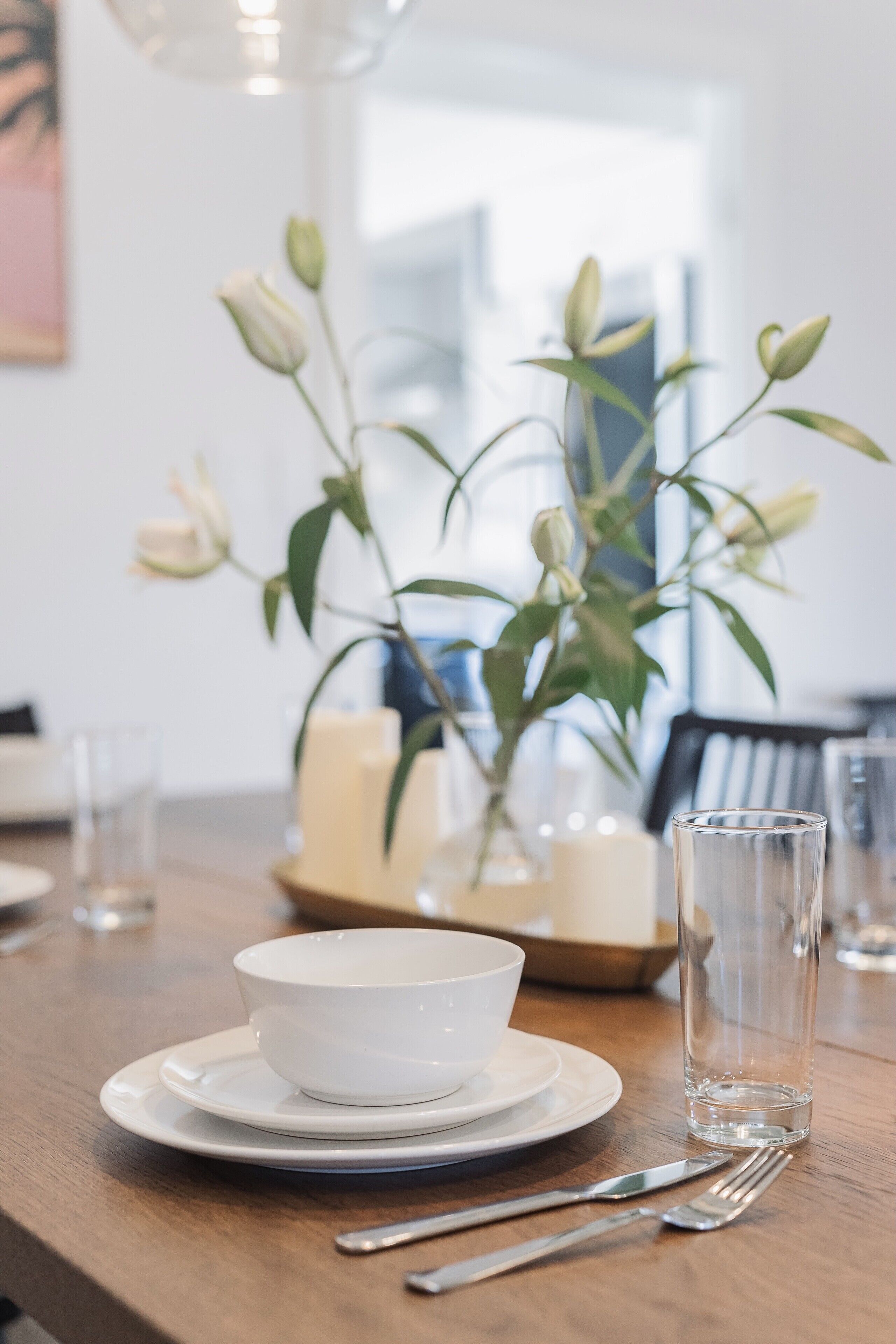A dining table with a white bowl, plates, cutlery, and a clear glass. There is a vase with green leaves and white buds in the background.
