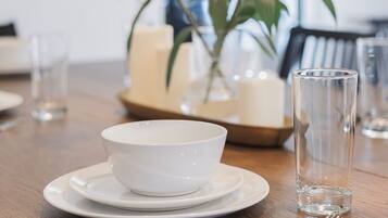 A dining table with a white bowl, plates, cutlery, and a clear glass. There is a vase with green leaves and white buds in the background.