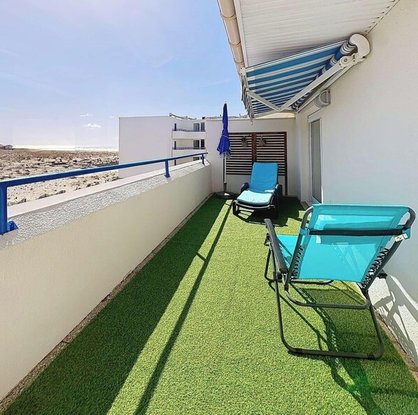 Un balcon avec du gazon synthétique, deux chaises longues, un parasol bleu et une vue sur la plage.