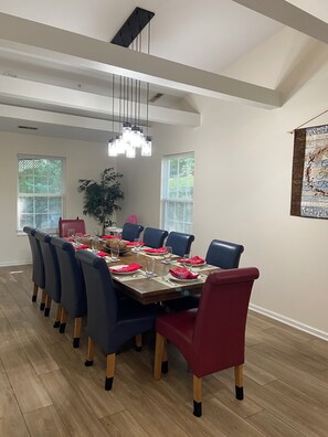 A dining area with a long table set for six, surrounded by blue and red chairs, under a modern chandelier.