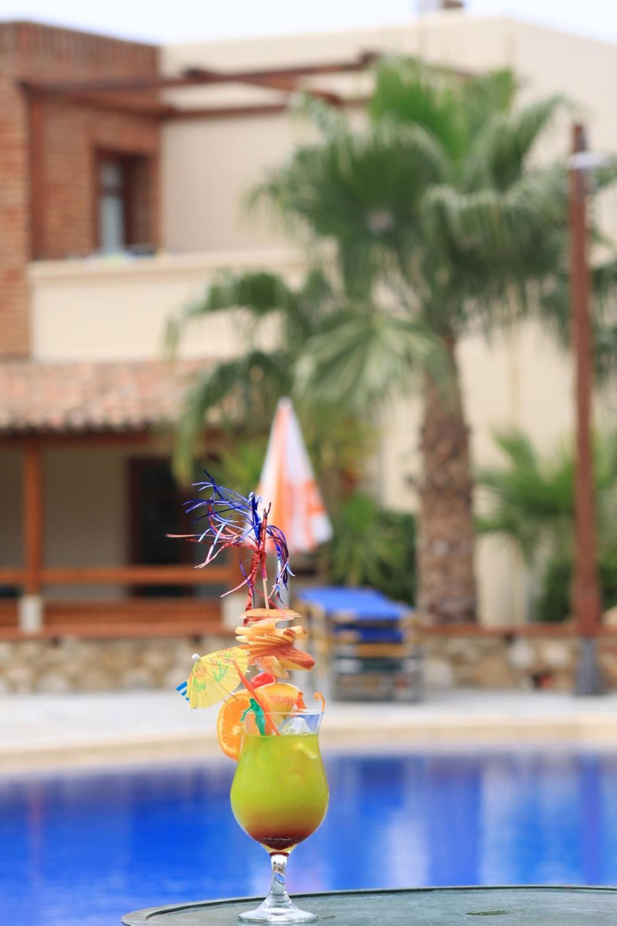 A cocktail on a table by a pool with a building and palm trees in the background.