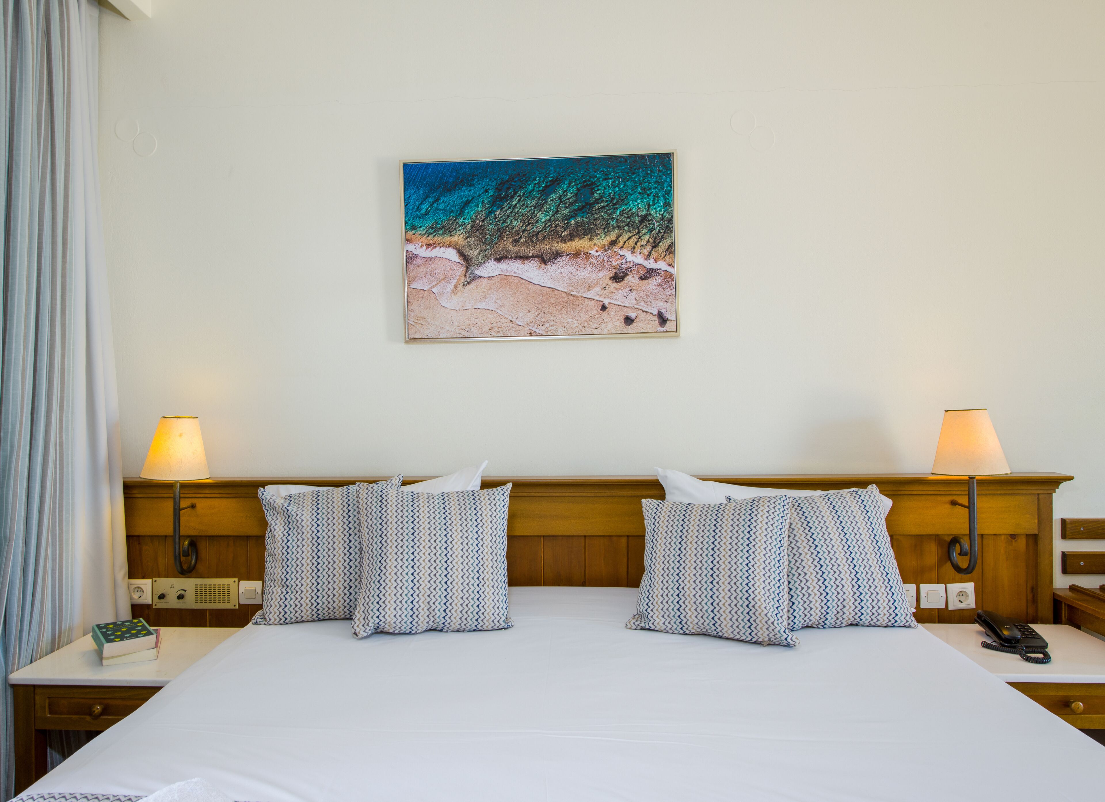 Une chambre d’hôtel avec un grand lit, deux tables de chevet avec des lampes, un tableau représentant une scène de plage accroché au mur, et un téléphone placé sur le côté droit du lit.