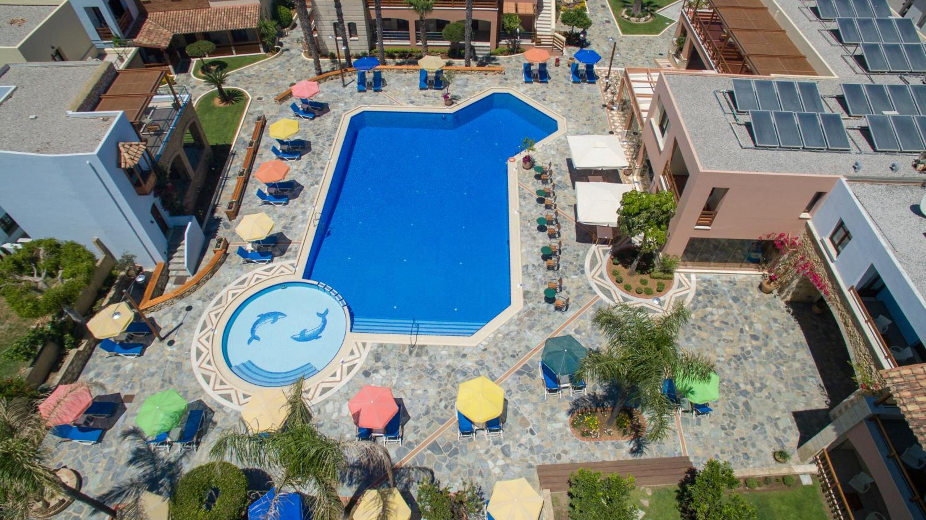 An aerial view of a resort with a central swimming pool surrounded by lounge chairs and umbrellas, with buildings and solar panels in the background.