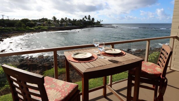 A wooden table with two plates and two glasses on a balcony overlooking the ocean. - Ocean Front Condo with AC, enjoy the ocean from your own private lanai. (Koloa)