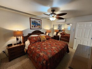 A bedroom with a bed, two bedside tables with lamps, a ceiling fan, a framed picture on the wall, and a door. - Ocean Front Condo with AC, enjoy the ocean from your own private lanai. (Koloa)