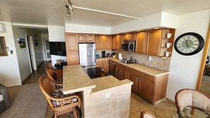 A kitchen with wooden cabinets, a stainless steel refrigerator, a microwave, and a coffee maker. There is a dining area with wicker chairs and a glass table. - Ocean Front Condo with AC, enjoy the ocean from your own private lanai. (Koloa)