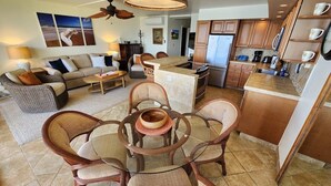 A spacious living area with a round wooden table, wicker chairs, and a sofa. A kitchen with wooden cabinets and a refrigerator is visible in the background. - Ocean Front Condo with AC, enjoy the ocean from your own private lanai. (Koloa)