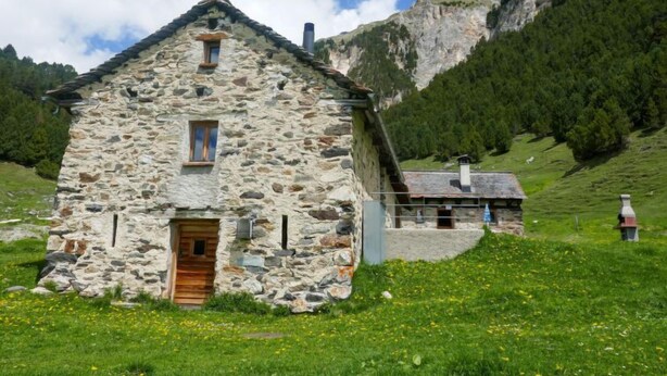 A stone house with a wooden door, set in a grassy field with a mountain in the background. - Rustico Dolomia by Interhome (Olivone)