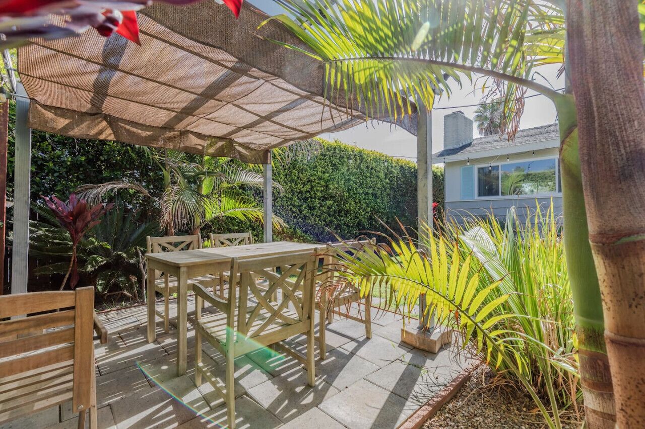 An outdoor patio with a wooden table and chairs, surrounded by various plants and a pergola providing shade.