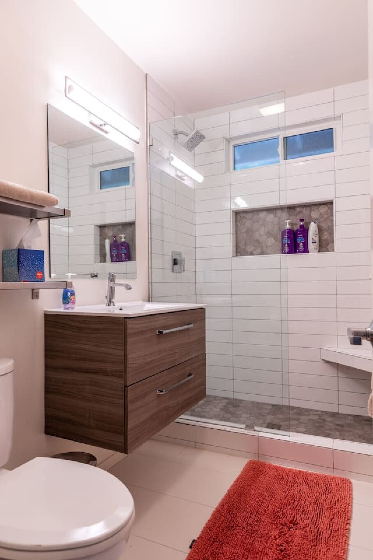 A modern bathroom with a white tiled shower, a wooden vanity, and a red rug.