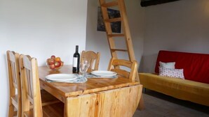 Ein Holztisch mit Stühlen, eine Weinflasche und eine Obstschale. Eine Holzleiter lehnt an der Wand. Im Hintergrund ist eine rote Couch mit einem weißen Kissen zu sehen.