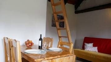 A wooden dining table with chairs, a wine bottle, and a fruit bowl. A wooden ladder is leaning against the wall. A red sofa with a white pillow is visible in the background.