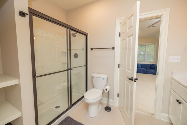 A modern bathroom with a glass shower enclosure, a toilet, and a visible hallway leading to a living area with a blue couch.