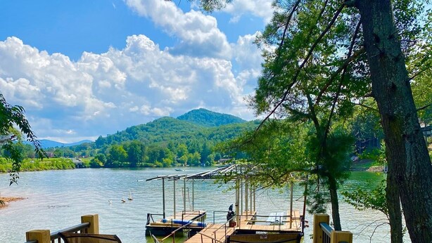 Una terraza de madera con sillas y mesas, con vistas a un lago, con un muelle y barcos al fondo.