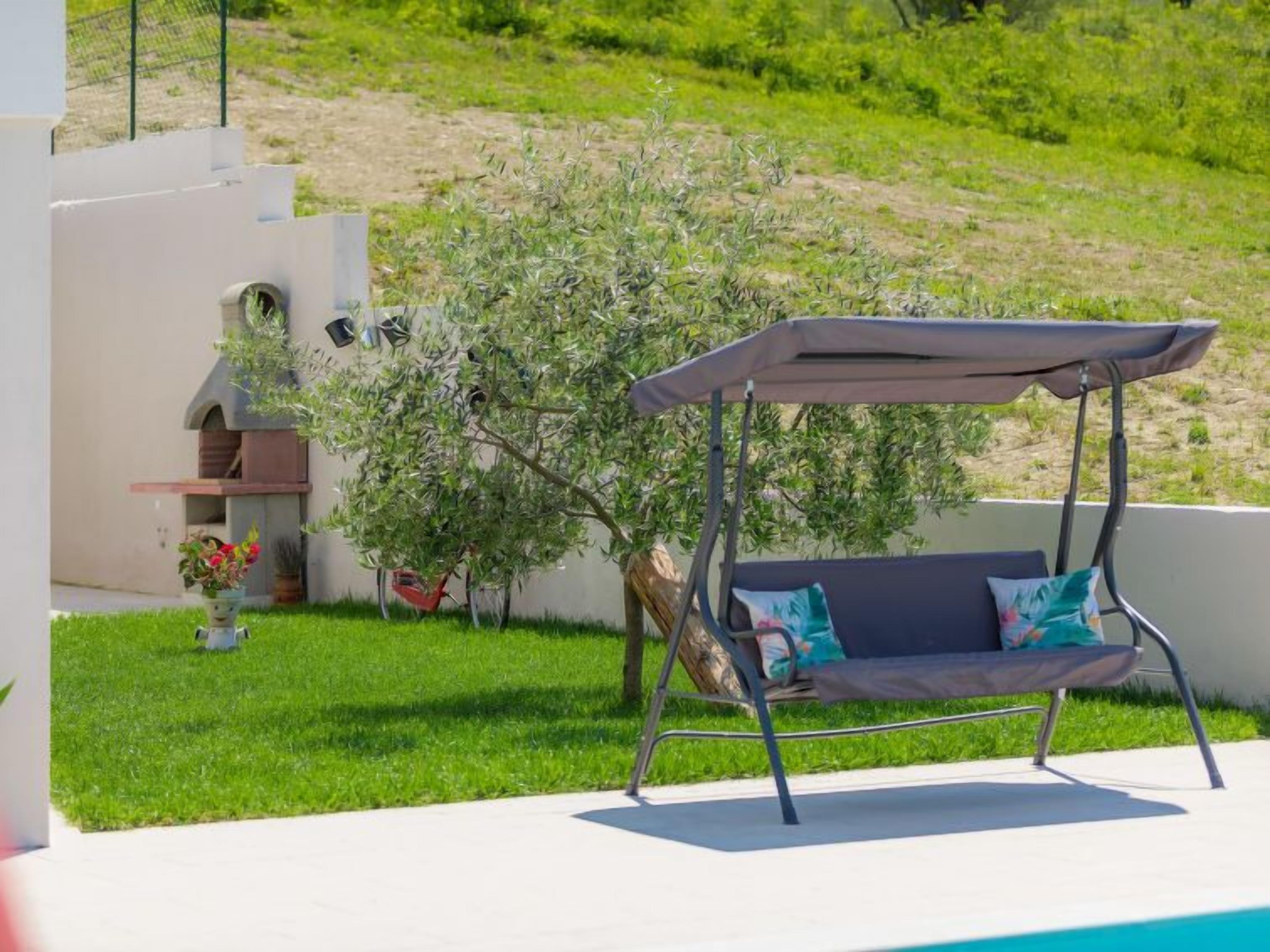 A poolside area with a swing chair, a small outdoor kitchen, and a view of a hillside.