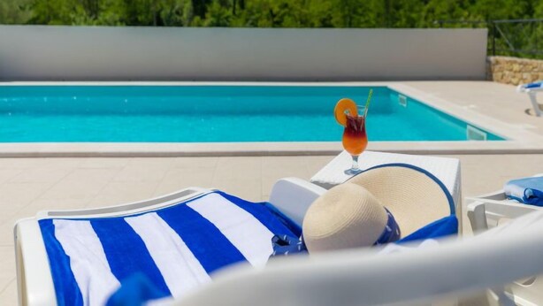 A poolside lounge chair with a striped towel and a sun hat, a glass of drink with an orange slice garnish, and a clear view of a swimming pool with a forested hillside in the background. - Montebello by Interhome (Motovun)