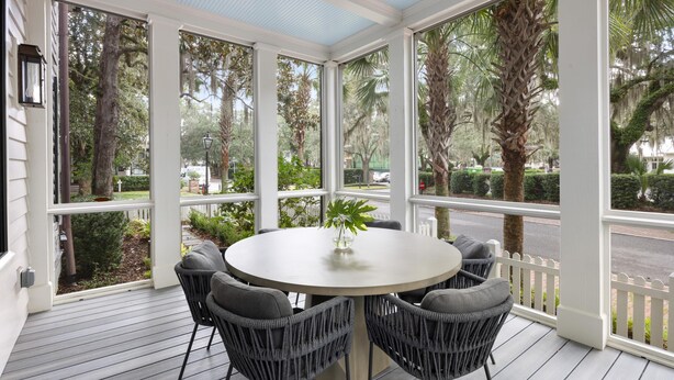 A covered patio with a round table and four chairs, large windows, and a view of trees and a street.