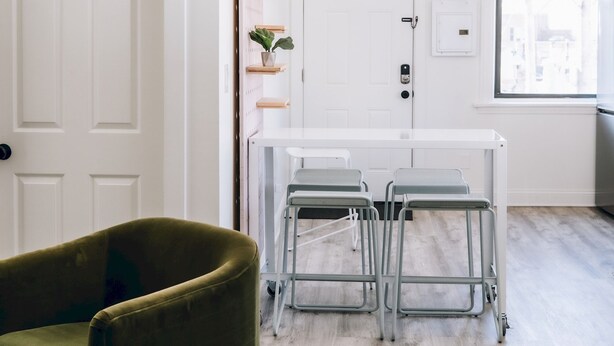 Une pièce moderne avec une porte blanche, un fauteuil vert, une table à manger avec des chaises en métal et une fenêtre donnant sur la ville.