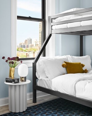 Ein Etagenbett mit weißer Bettwäsche und einem senfgelben Kissen, ein kleiner Tisch mit einem Blumenstrauß und ein Blick auf eine Stadtlandschaft durch ein Fenster.